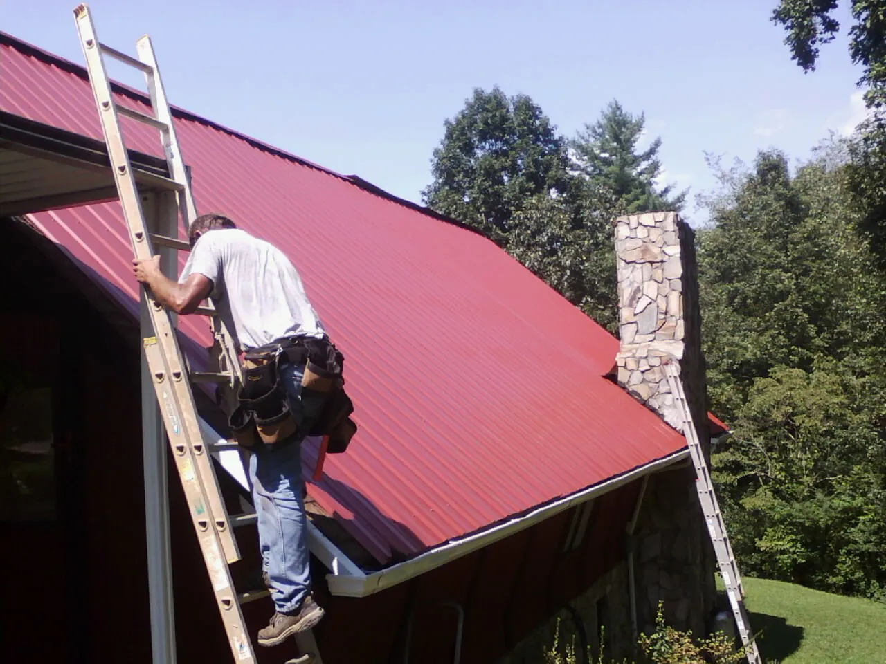 Roofer on a ladder beside a red metal roof and stone chimney in Greeley